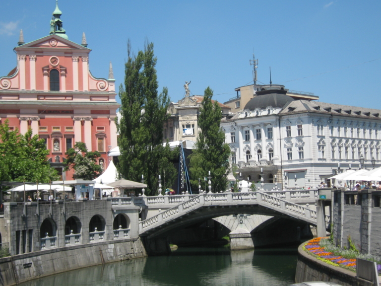 A stone bridge crosses a canal, bordered by historic pink and white buildings with trees and outdoor seating on a sunny day.