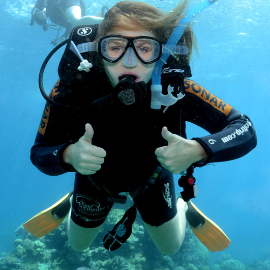A scuba diver underwater gives two thumbs up, wearing a wetsuit, mask, and fins, with coral visible in the background.