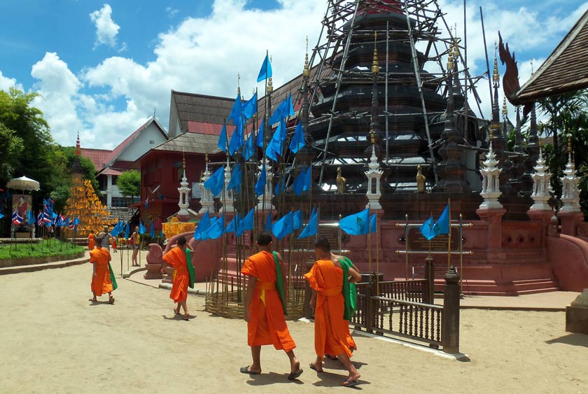 Four Buddhist monks in orange robes walk past a temple adorned with blue flags and scaffolding, under a partly cloudy sky.