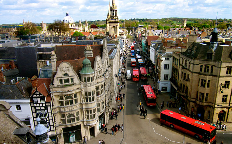 A busy street scene in Oxford, England, featuring historic buildings, pedestrians, and several red double-decker buses under a partly cloudy sky.