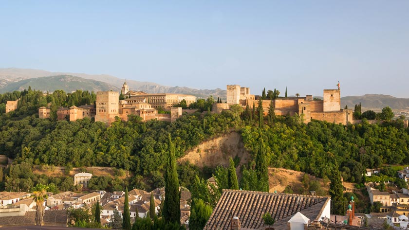 The Alhambra, a historic fortress and palace complex, sits on a lush, green hill overlooking the city of Granada with mountains in the background.