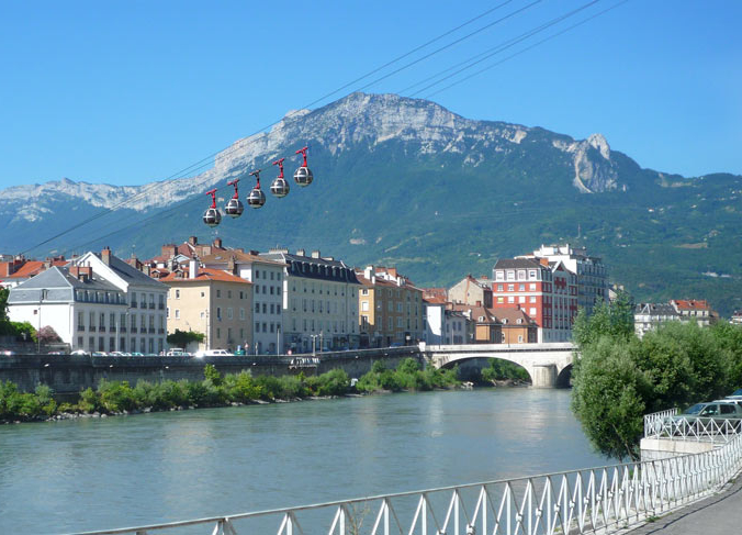 A river runs through a city with colorful buildings, a stone bridge, and cable cars overhead; mountains rise in the background under a clear blue sky.