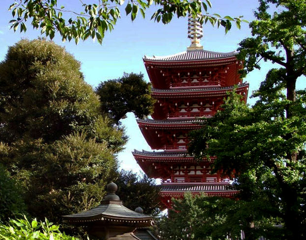 A red multi-tiered pagoda partially obscured by trees and greenery under a blue sky.