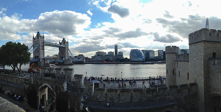 A panoramic view of the River Thames with Tower Bridge on the left, modern buildings across the water, and stone walls of the Tower of London in the foreground.
