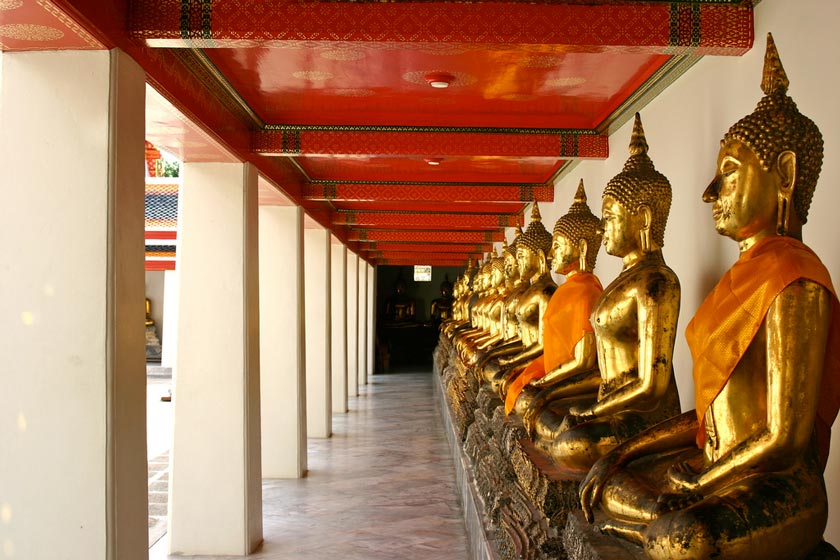 Row of gold Buddha statues draped in orange cloth, seated along a corridor with red ceiling and white columns in a temple.