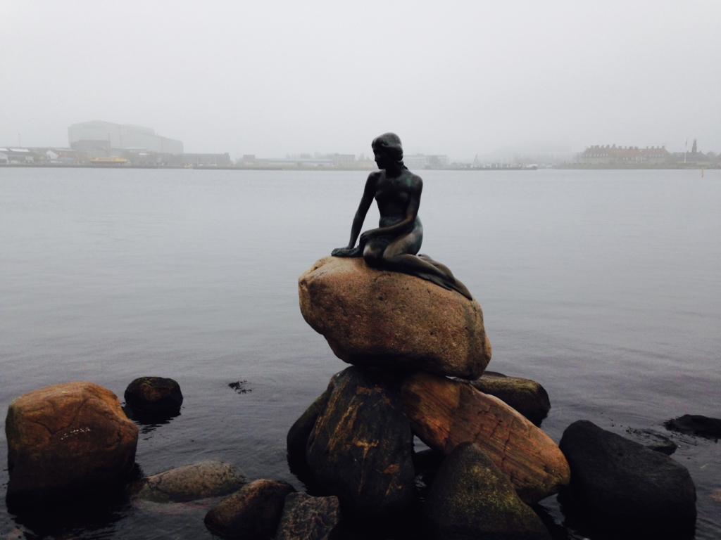 Bronze statue of a mermaid sitting on a rock by the water, with a foggy coastline and buildings in the background.