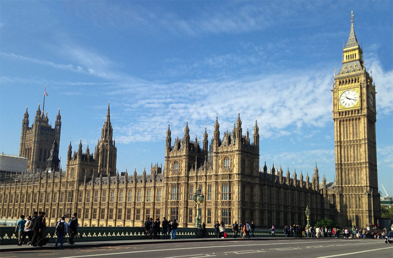 The Palace of Westminster with the Elizabeth Tower, known as Big Ben, under a blue sky with people walking on the street in front.