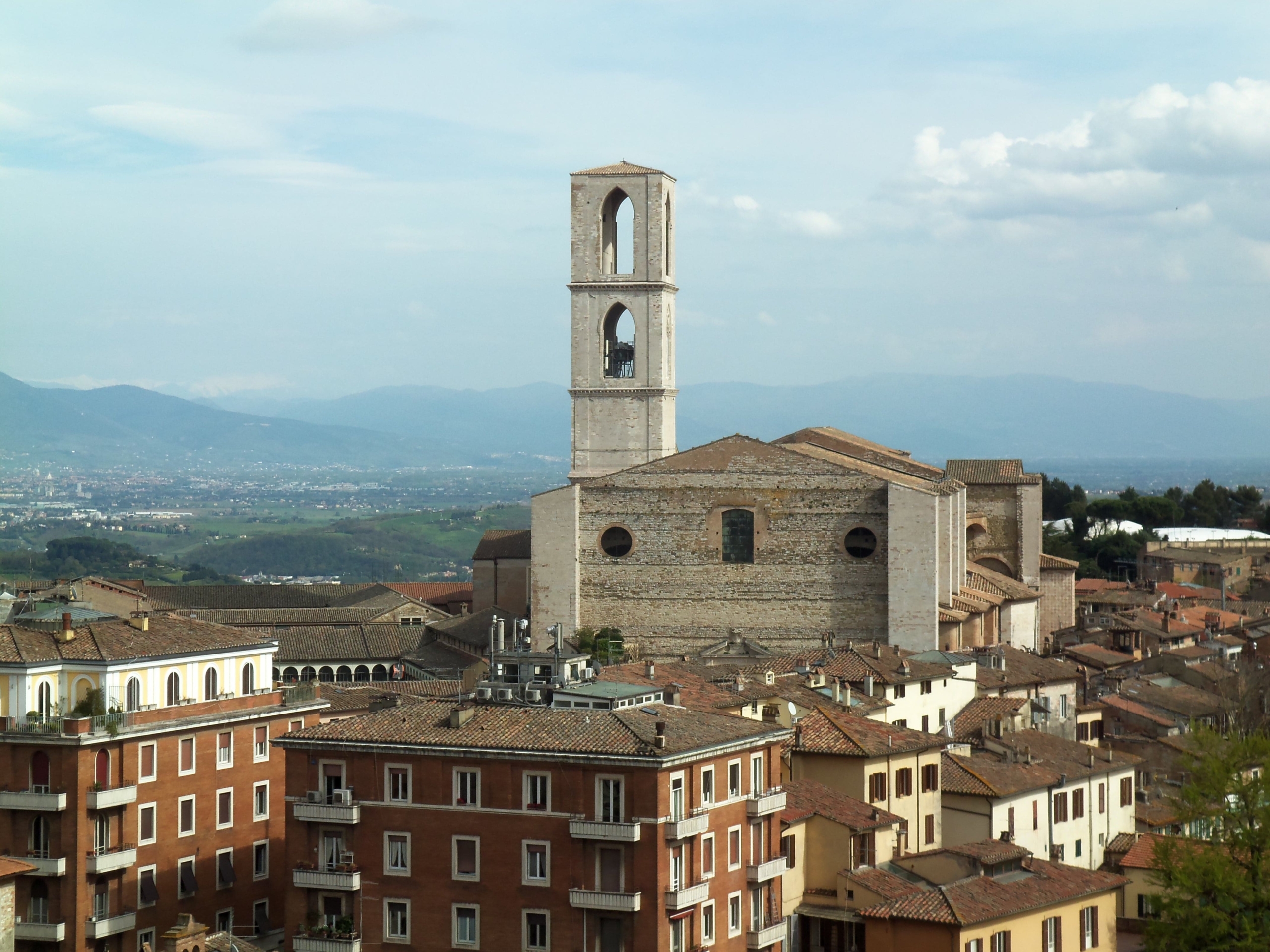 A large stone church with a prominent rectangular bell tower stands among red-roofed buildings in a historic town with rolling hills and mountains in the background.
