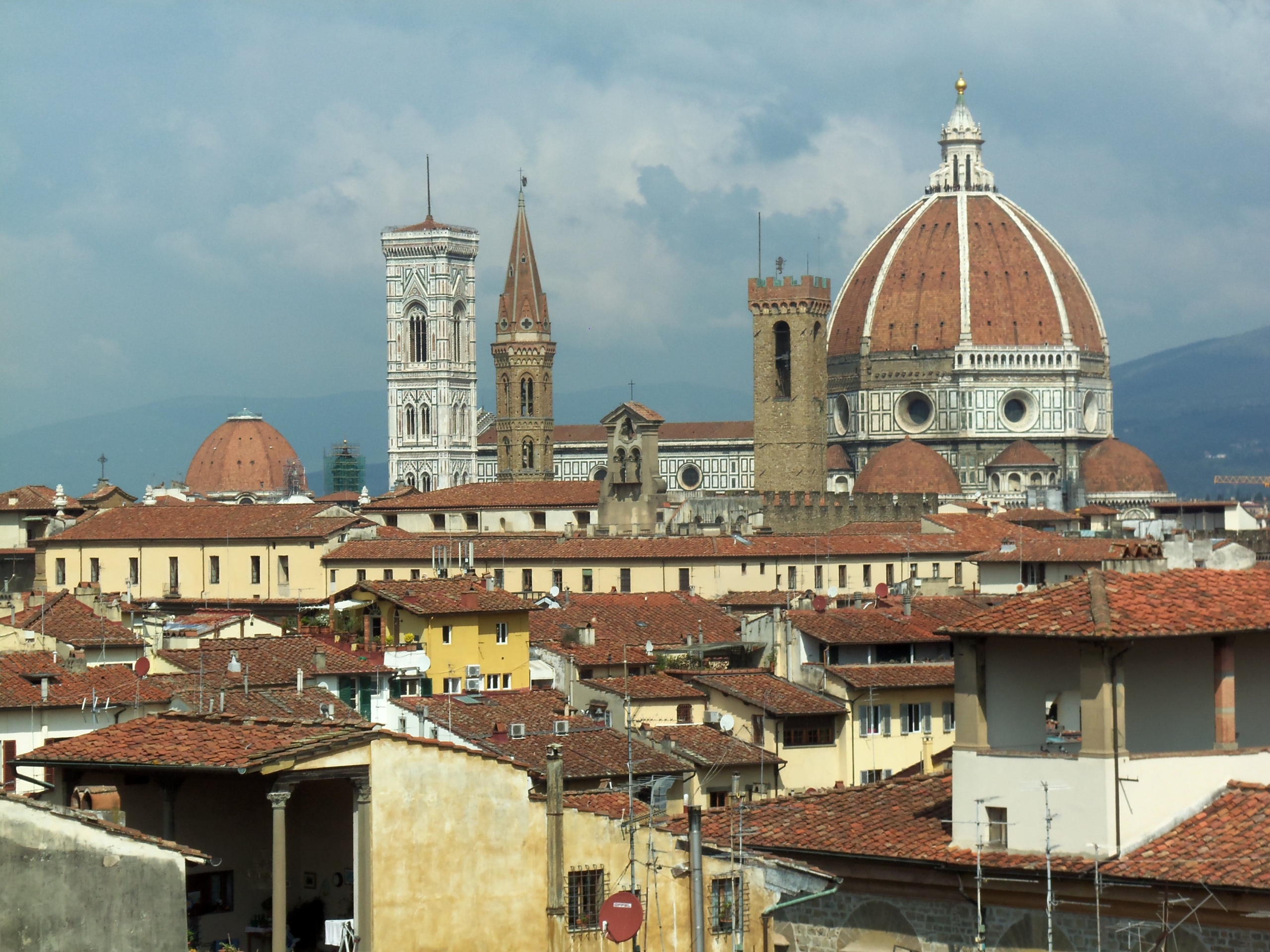 Florence cityscape with terracotta rooftops, featuring the Florence Cathedral, its dome, bell tower, and surrounding historic buildings under a partly cloudy sky.