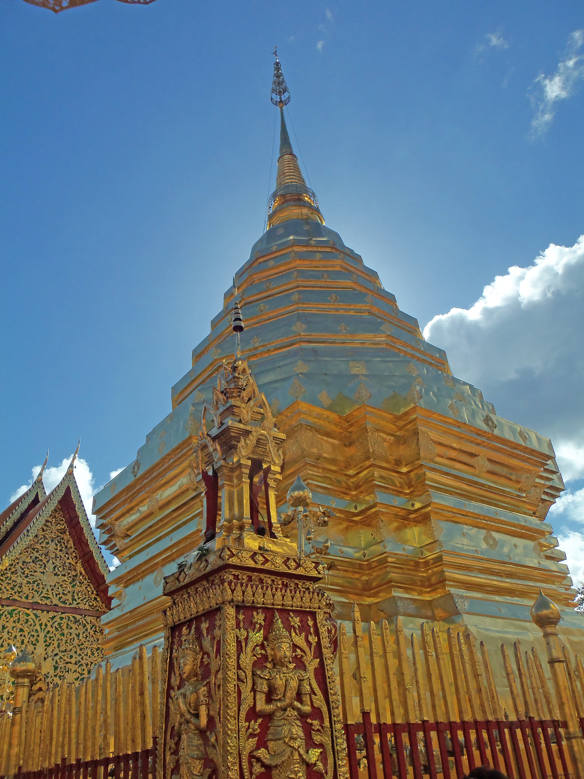 A golden Buddhist stupa with ornate carvings and statues stands against a blue sky with clouds, surrounded by a decorative fence.