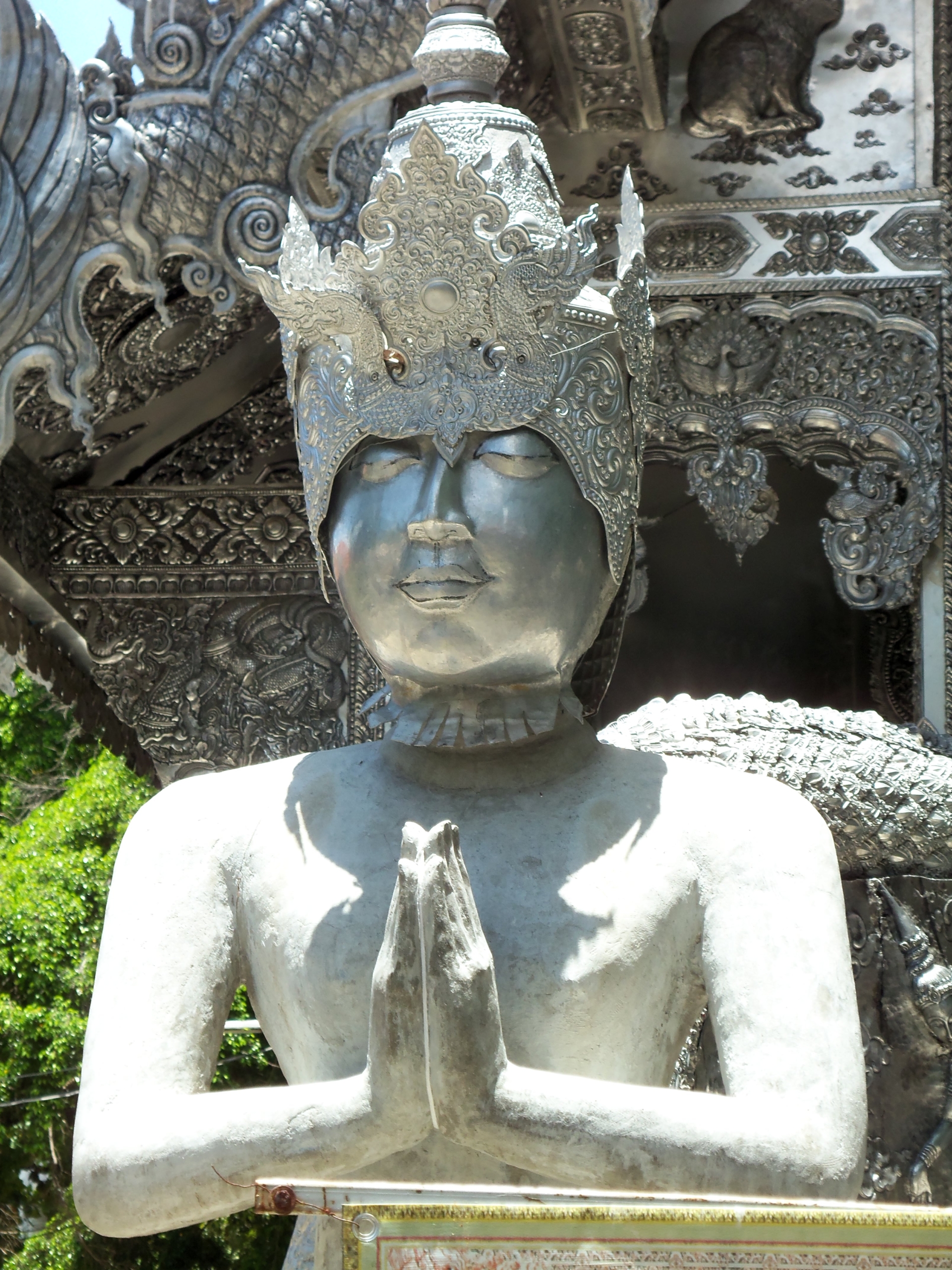 A stone Buddhist statue in a prayer pose wearing an ornate silver crown, set against the intricate silver facade of a Thai temple.