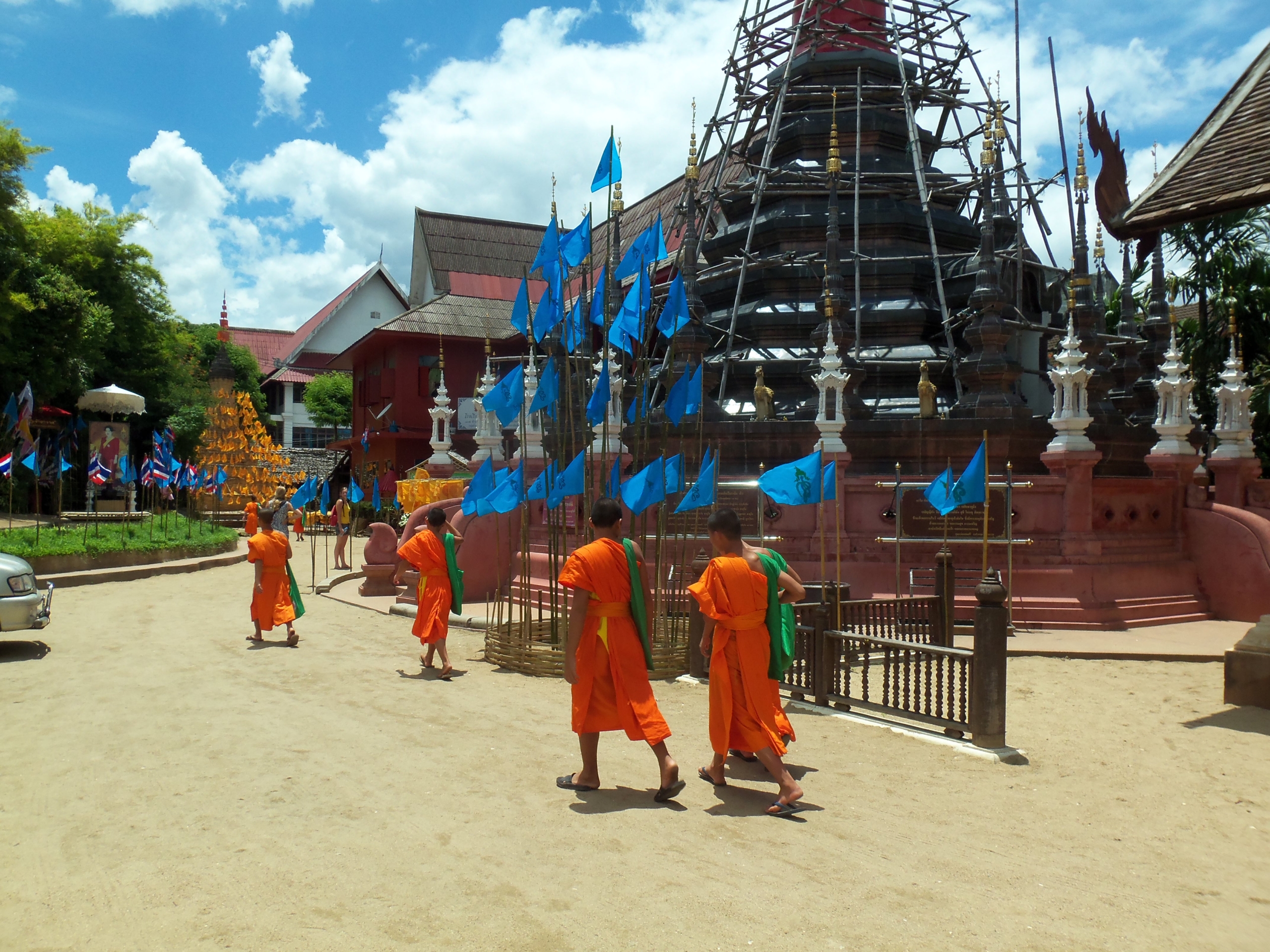Four Buddhist monks in orange robes walk past a temple adorned with blue flags and scaffolding under a bright, sunny sky.