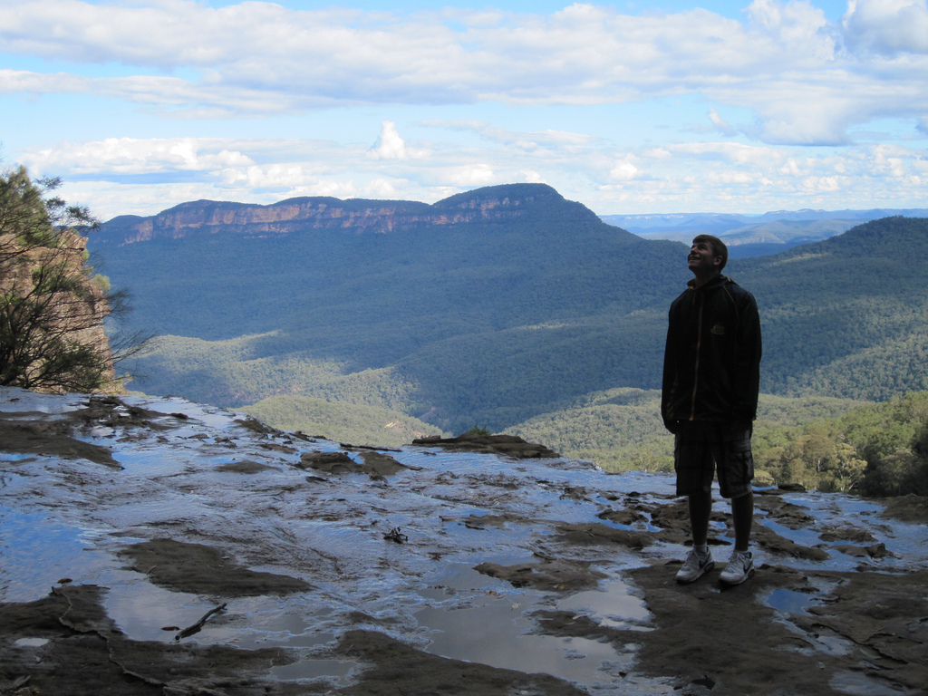 A person stands on a rocky, wet surface overlooking a vast forested valley and distant mountains under a partly cloudy sky.