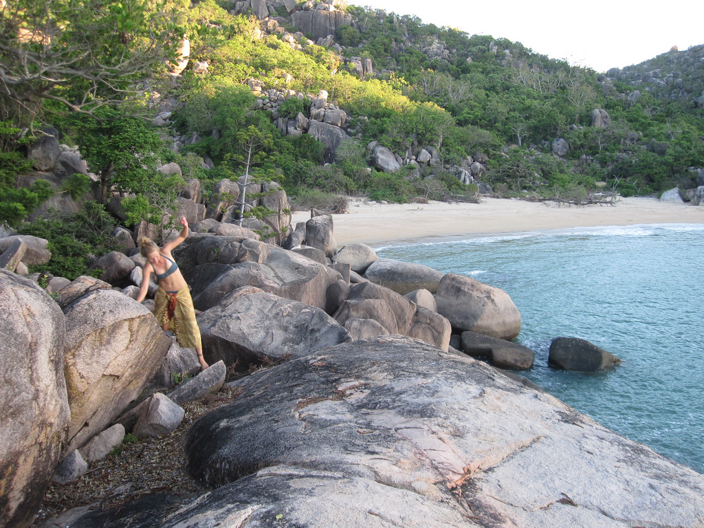 A person climbs over large rocks near a secluded beach with dense greenery and hills in the background.