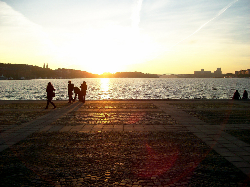 People walking along a paved waterfront promenade at sunset, with a river and distant bridge under a clear sky.