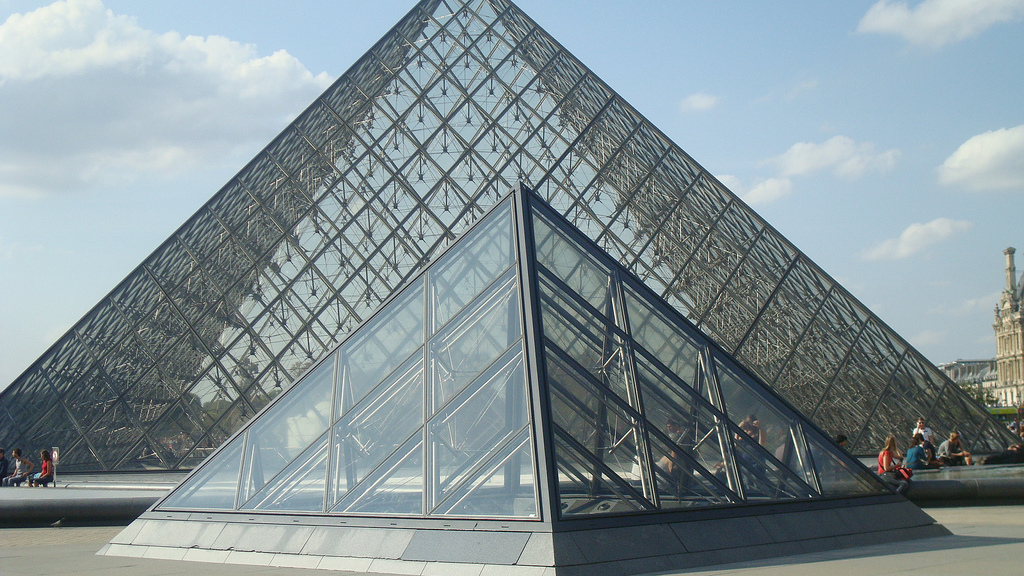 Glass pyramid structures at the Louvre Museum in Paris, with the larger main pyramid and a smaller one in the foreground under a partly cloudy sky.
