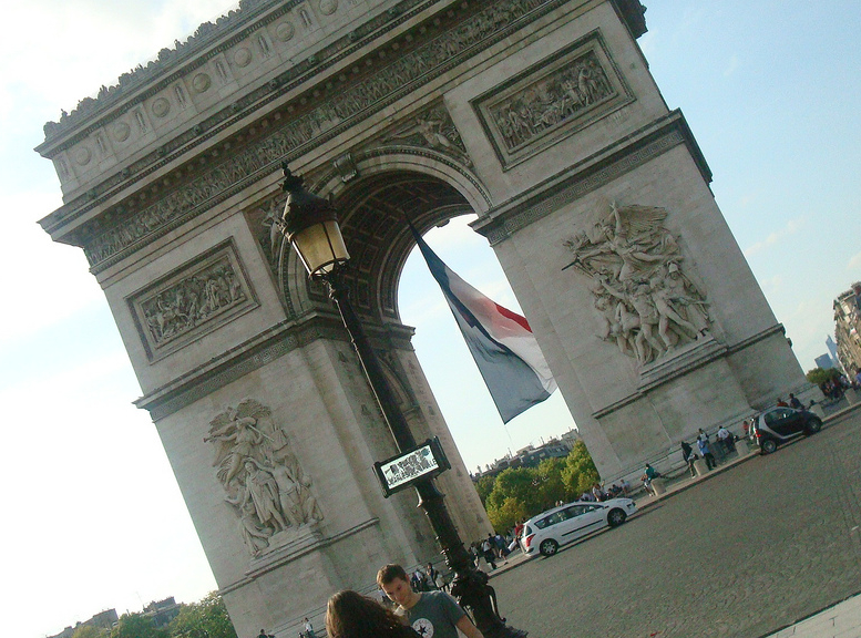 The Arc de Triomphe in Paris with a large French flag hanging beneath the arch, cars and people visible nearby, photo taken at an angle.