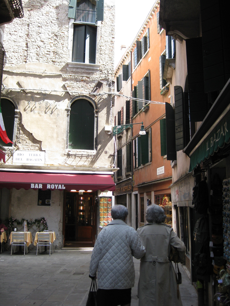 Two elderly people walk arm-in-arm down a narrow European street lined with shops, restaurants, and old buildings with green shutters.