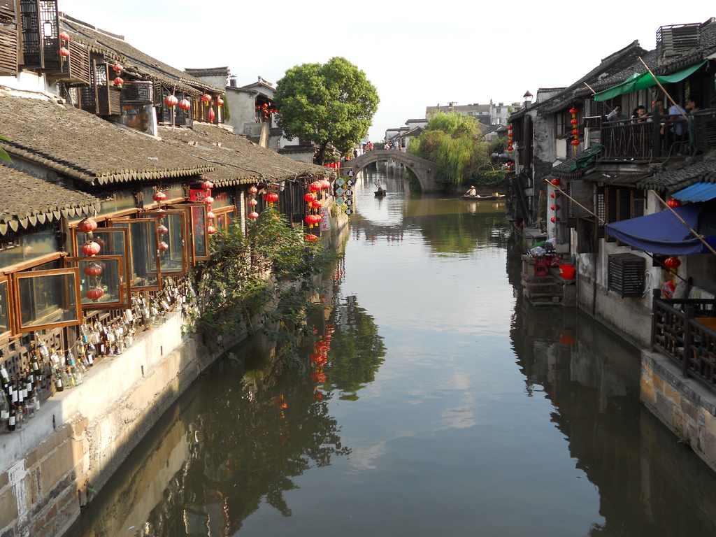 Traditional Chinese water town with stone buildings, red lanterns, open windows, and a canal leading to a stone arch bridge under a clear sky.