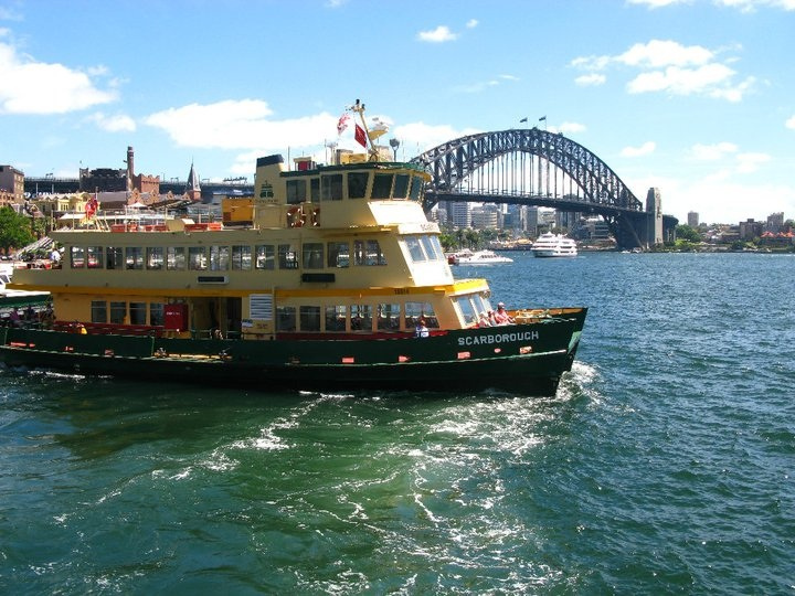 A yellow and green ferry labeled "Scarborough" sails on the water with Sydney Harbour Bridge and city buildings visible in the background under a clear sky.