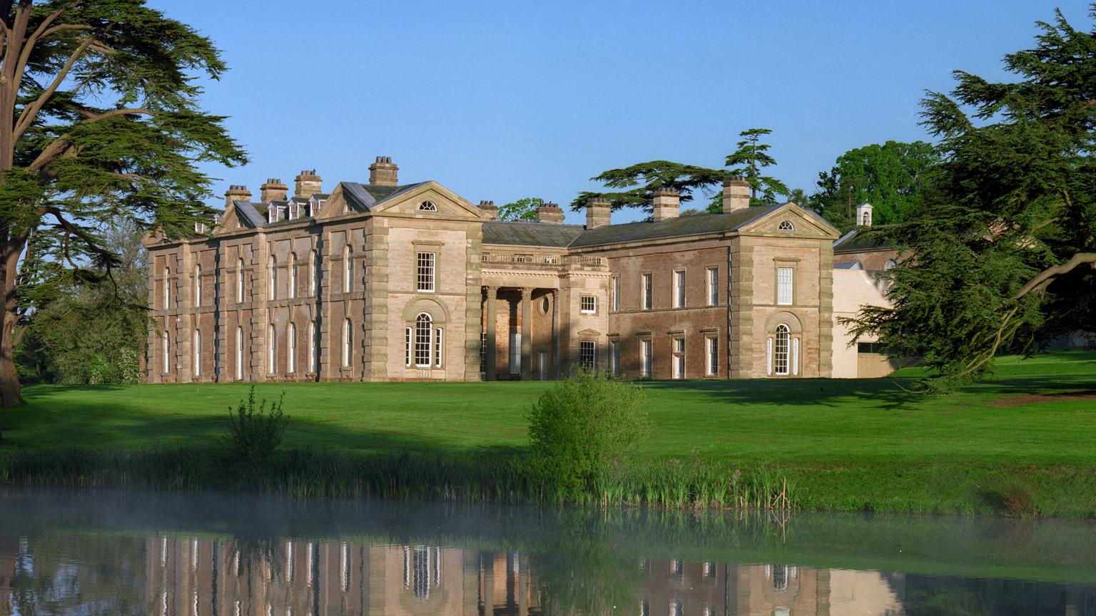 Large historic mansion with classical columns, surrounded by green lawn, mature trees, and a reflective pond in the foreground under a clear blue sky.