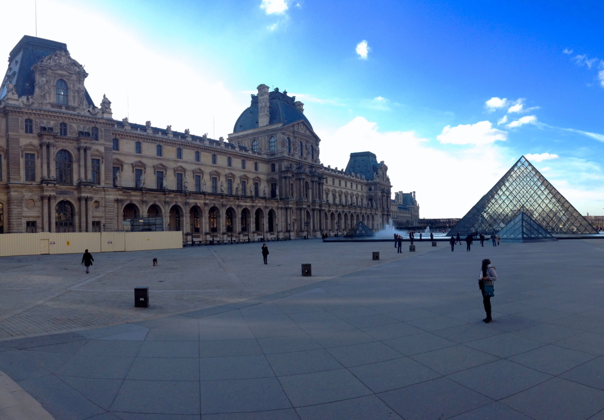 The Louvre Museum in Paris with its glass pyramid entrance, people walking around the large open courtyard under a blue sky.