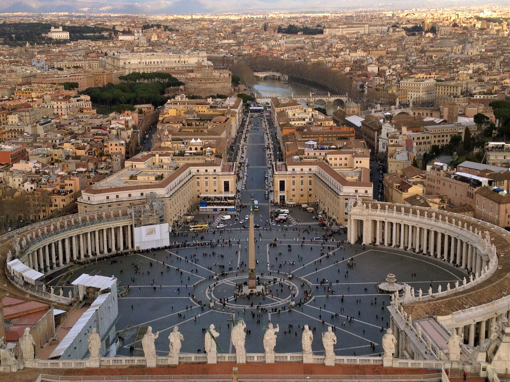 Aerial view of St. Peter’s Square and the surrounding colonnades in Vatican City, with the city of Rome and the Tiber River in the background.