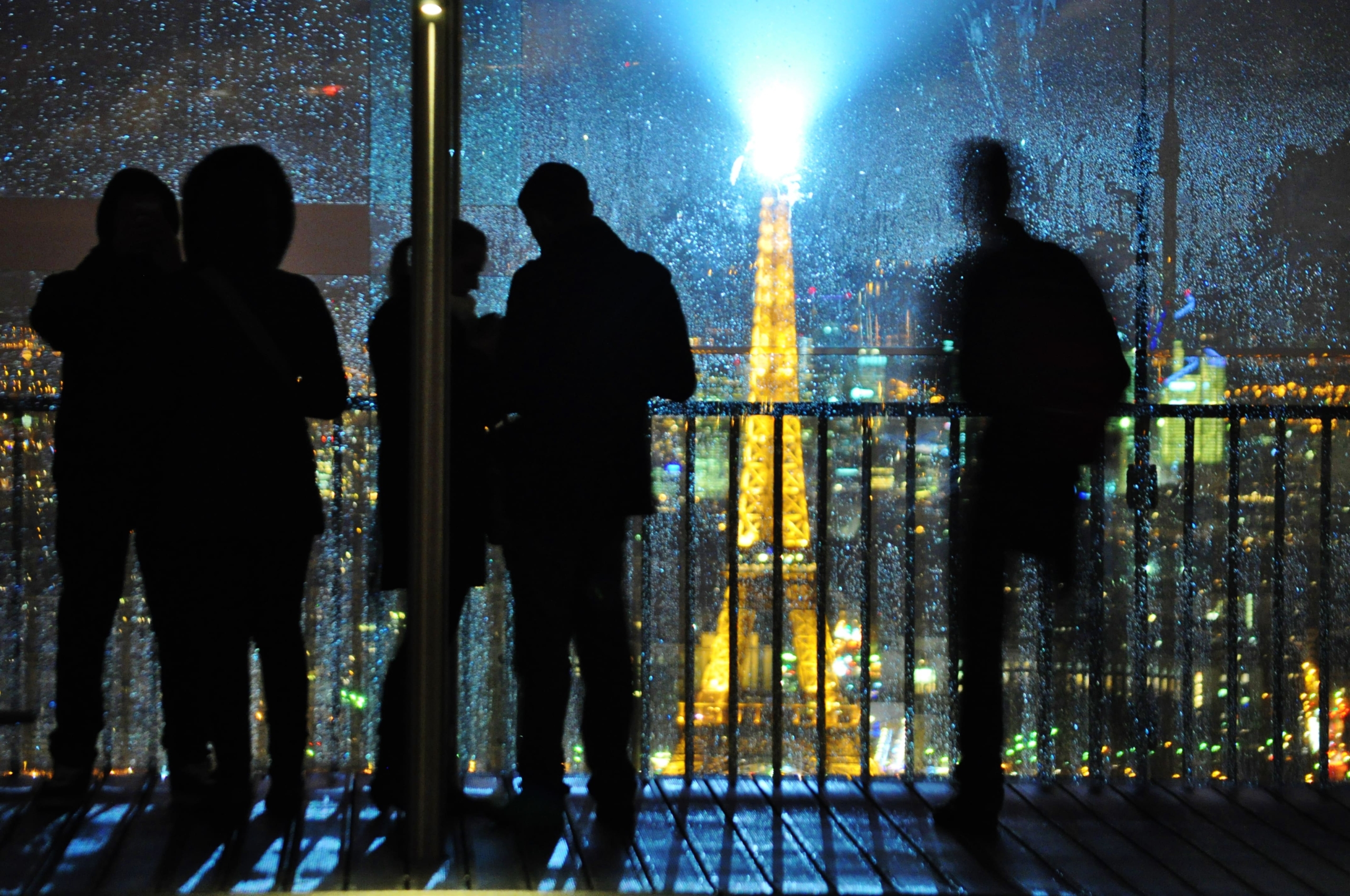 Silhouettes of people stand on a balcony overlooking a cityscape at night, with the illuminated Eiffel Tower visible through a rain-speckled window.