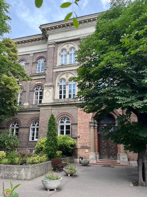 A three-story brick building with arched windows and a large wooden door, surrounded by trees, planters, and a bench on a paved walkway.