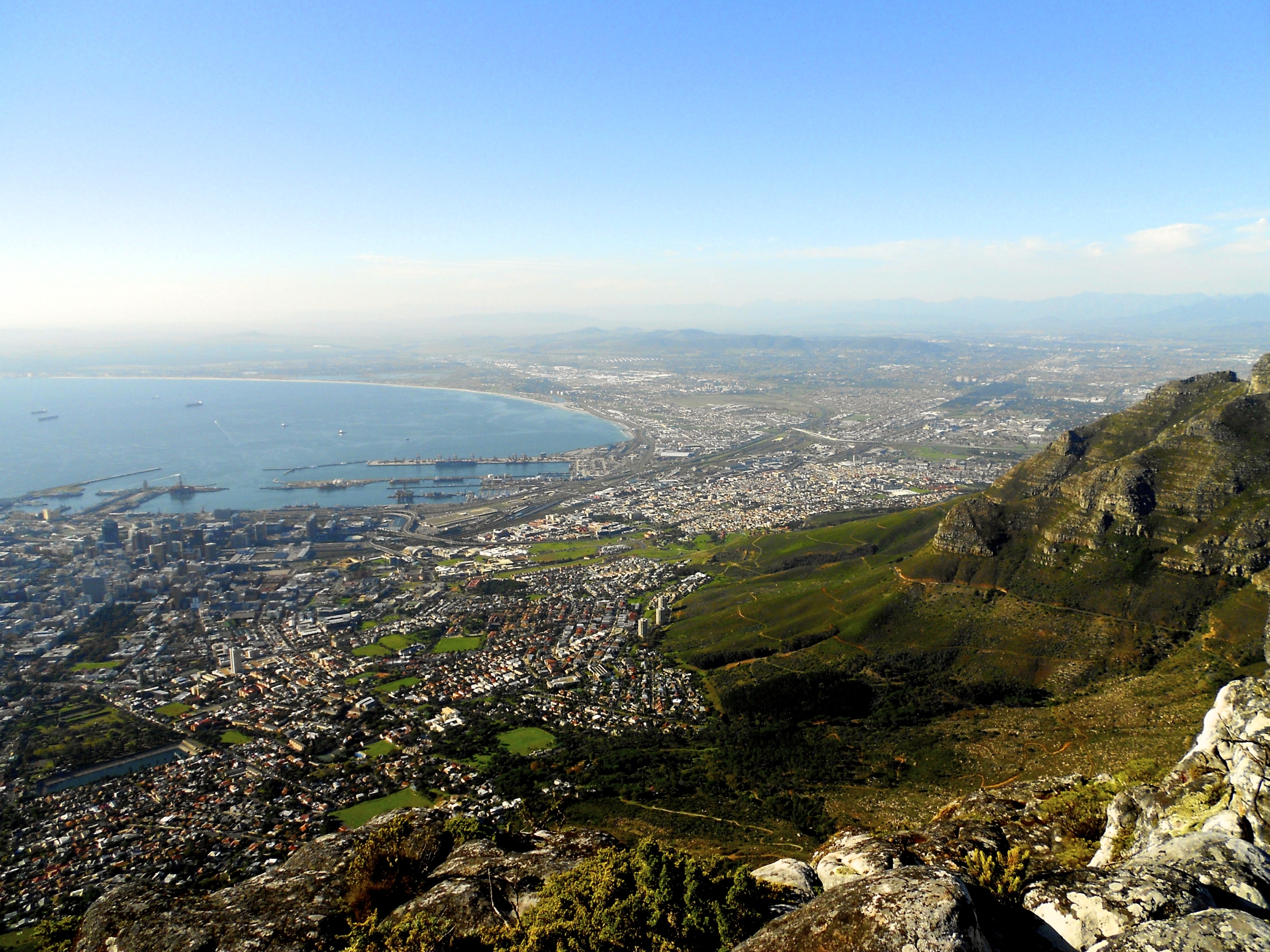 View from a mountain overlooking a coastal city, with urban areas, green spaces, and the sea visible under a clear blue sky.
