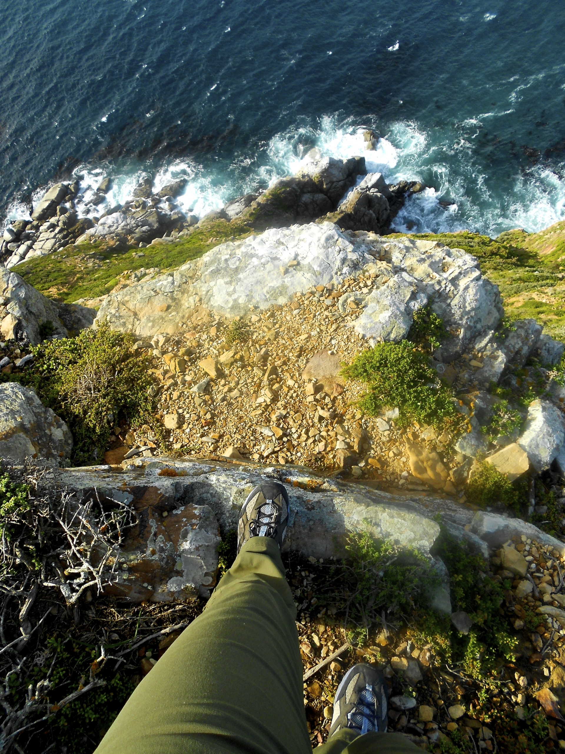 Person standing at the edge of a rocky cliff above the ocean, with waves crashing against rocks below, seen from a first-person perspective.