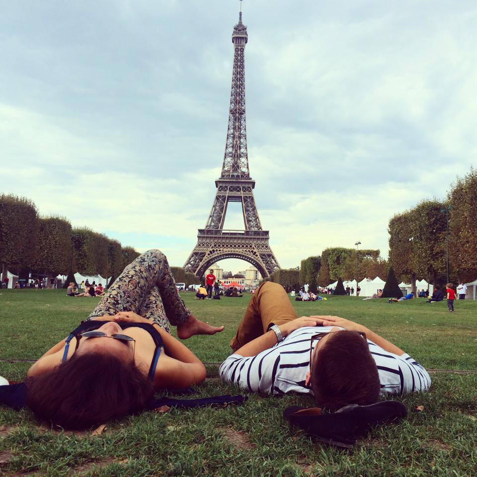Two people lie on the grass facing the Eiffel Tower in Paris, with trees and other visitors visible in the background under a cloudy sky.