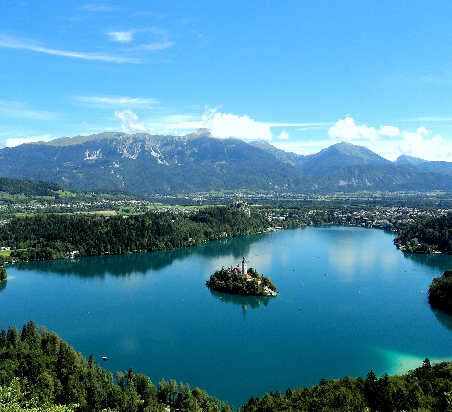 A clear blue lake with a small island in the center, surrounded by green hills and mountains under a bright blue sky.