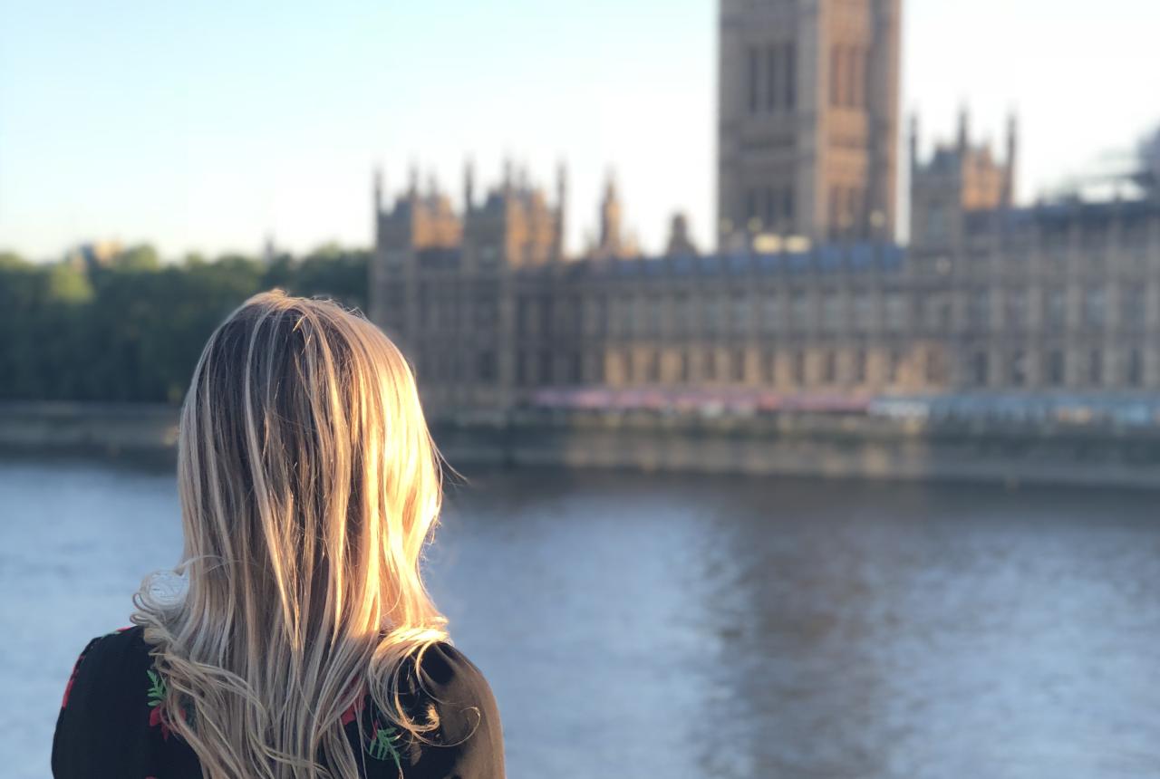 A person with long blonde hair stands by the river, facing the Palace of Westminster in London on a sunny day.