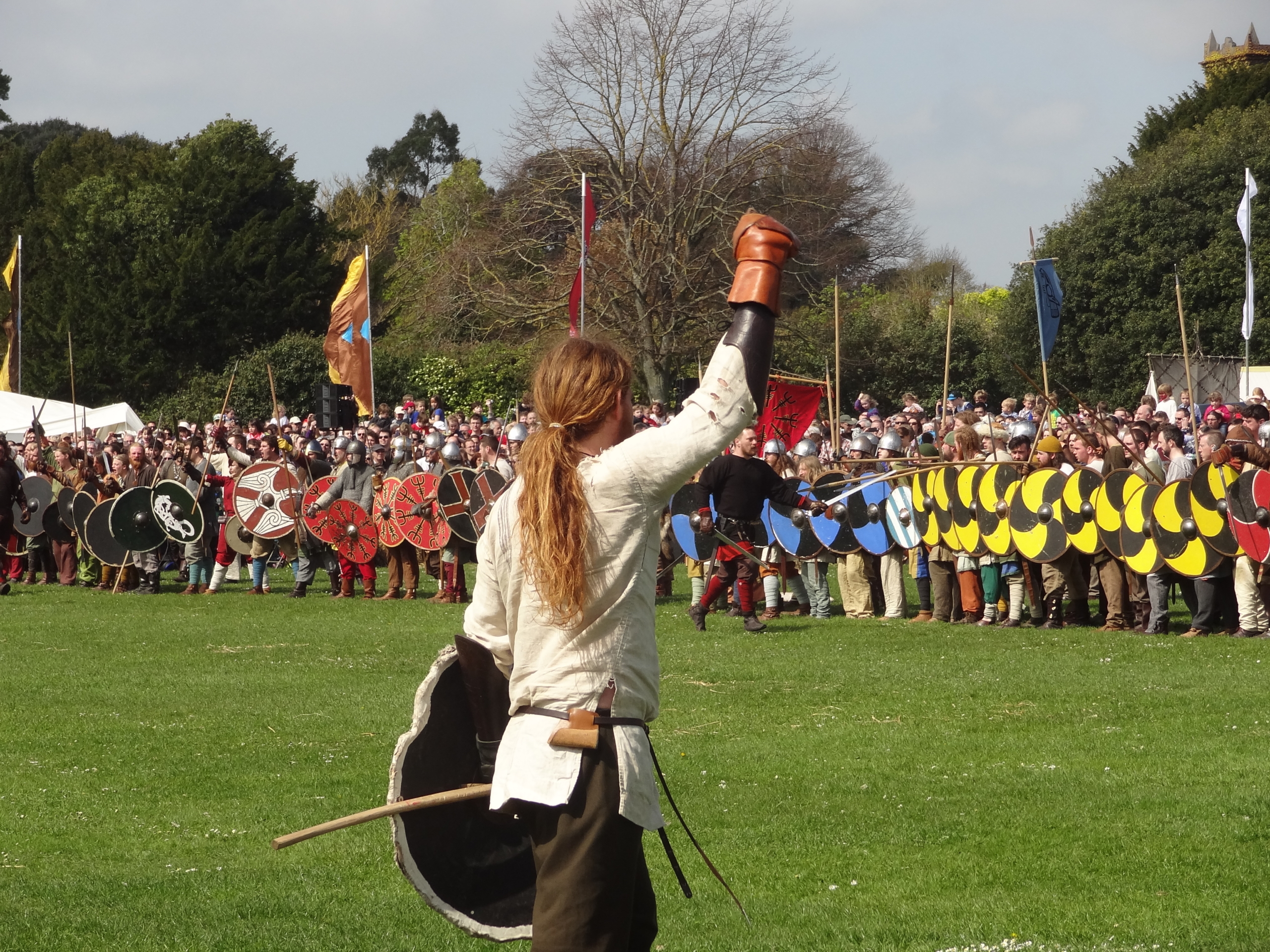 A person in historical costume raises a gloved fist in front of a large group of reenactors with shields lined up outdoors.