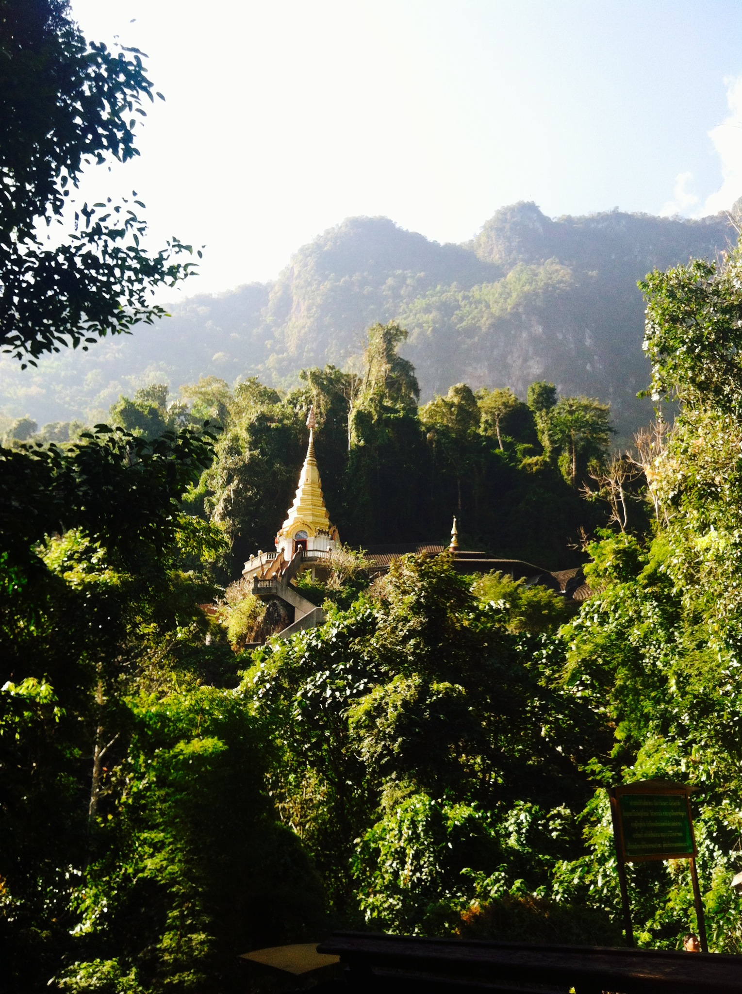 A golden stupa surrounded by dense green forest with mountains in the background under a bright sky.