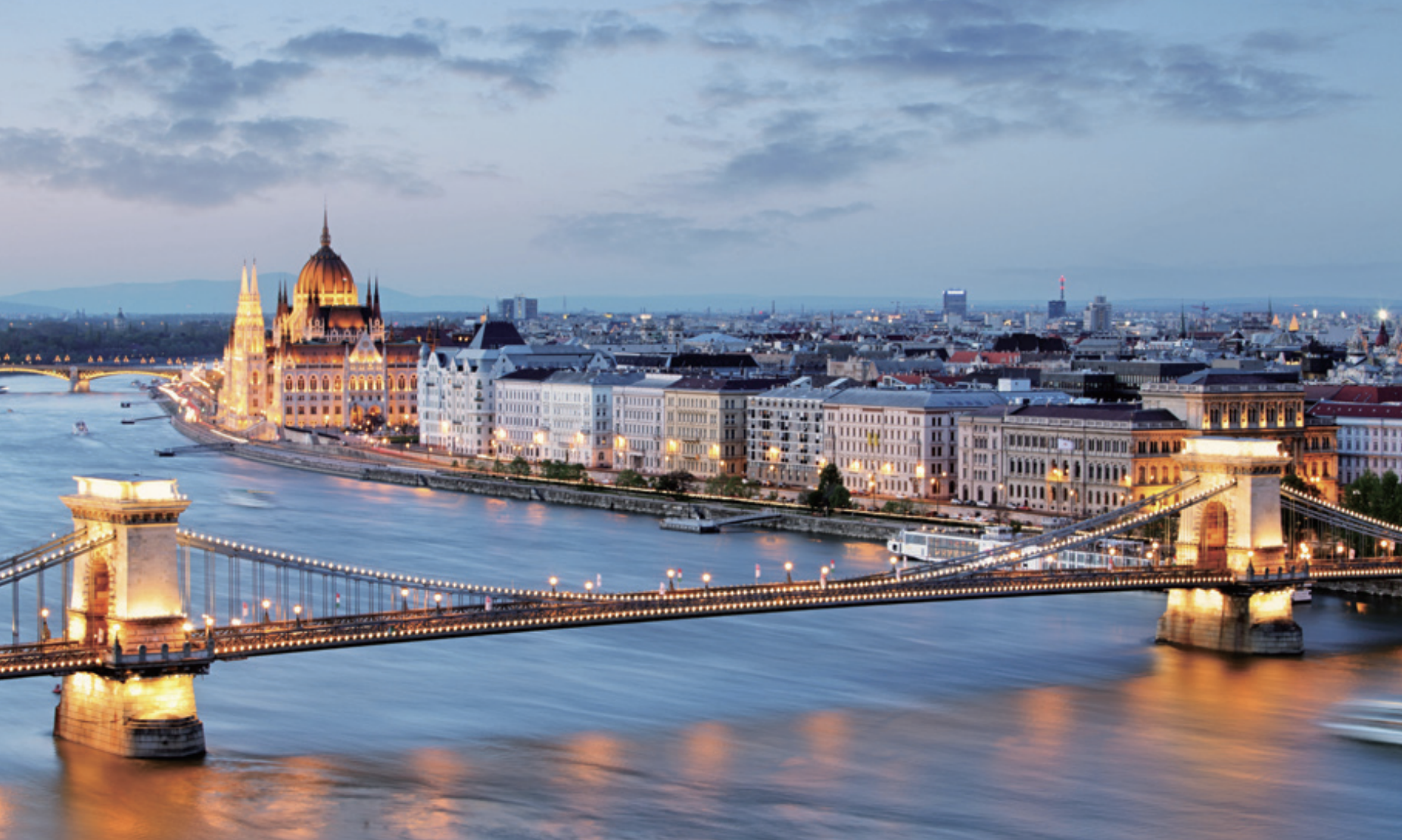 A cityscape view of Budapest at dusk, showing the Chain Bridge over the Danube River and the illuminated Hungarian Parliament building.