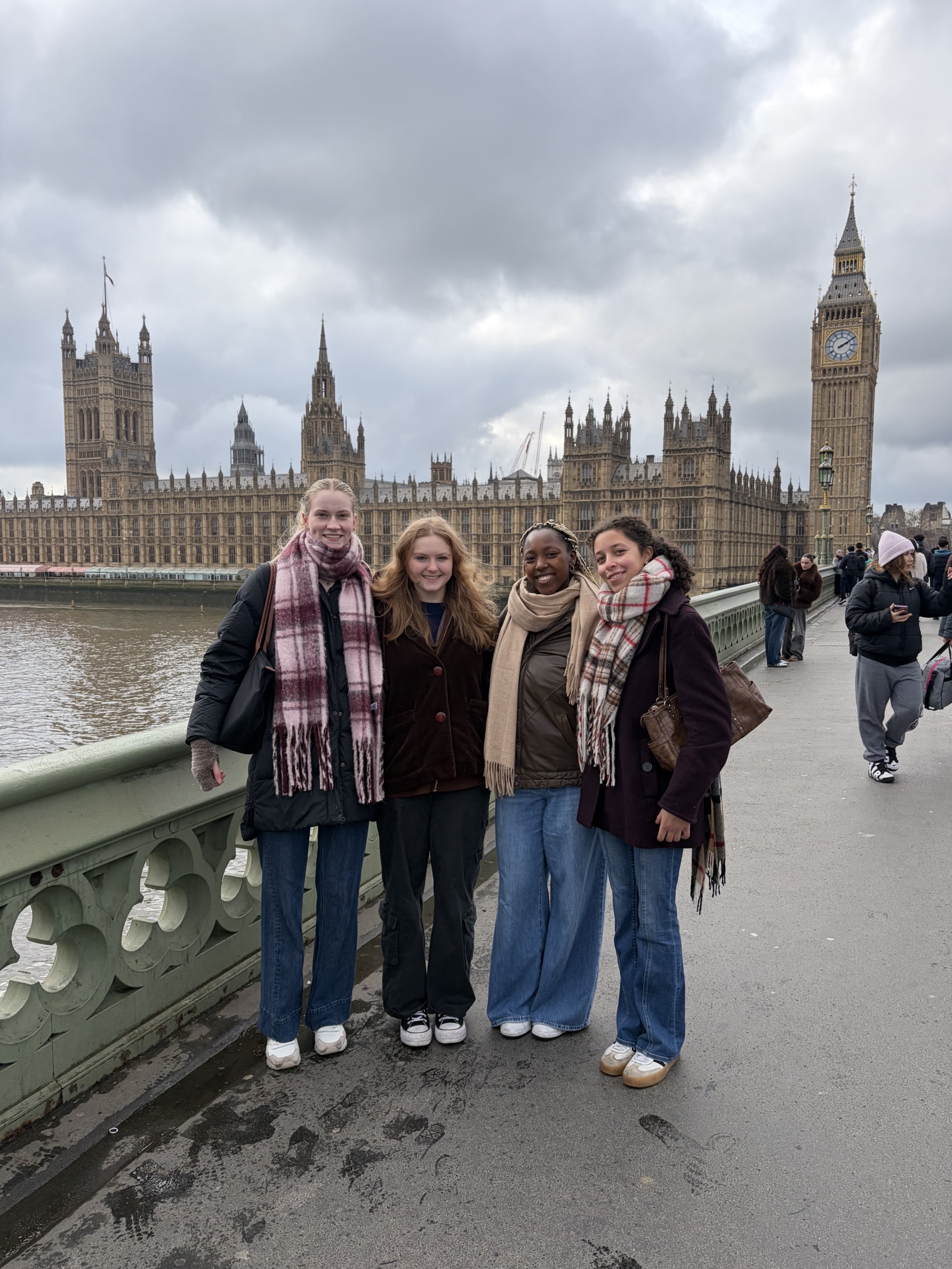 Four women stand side by side on Westminster Bridge in London, with the Houses of Parliament and Big Ben visible in the background on a cloudy day.