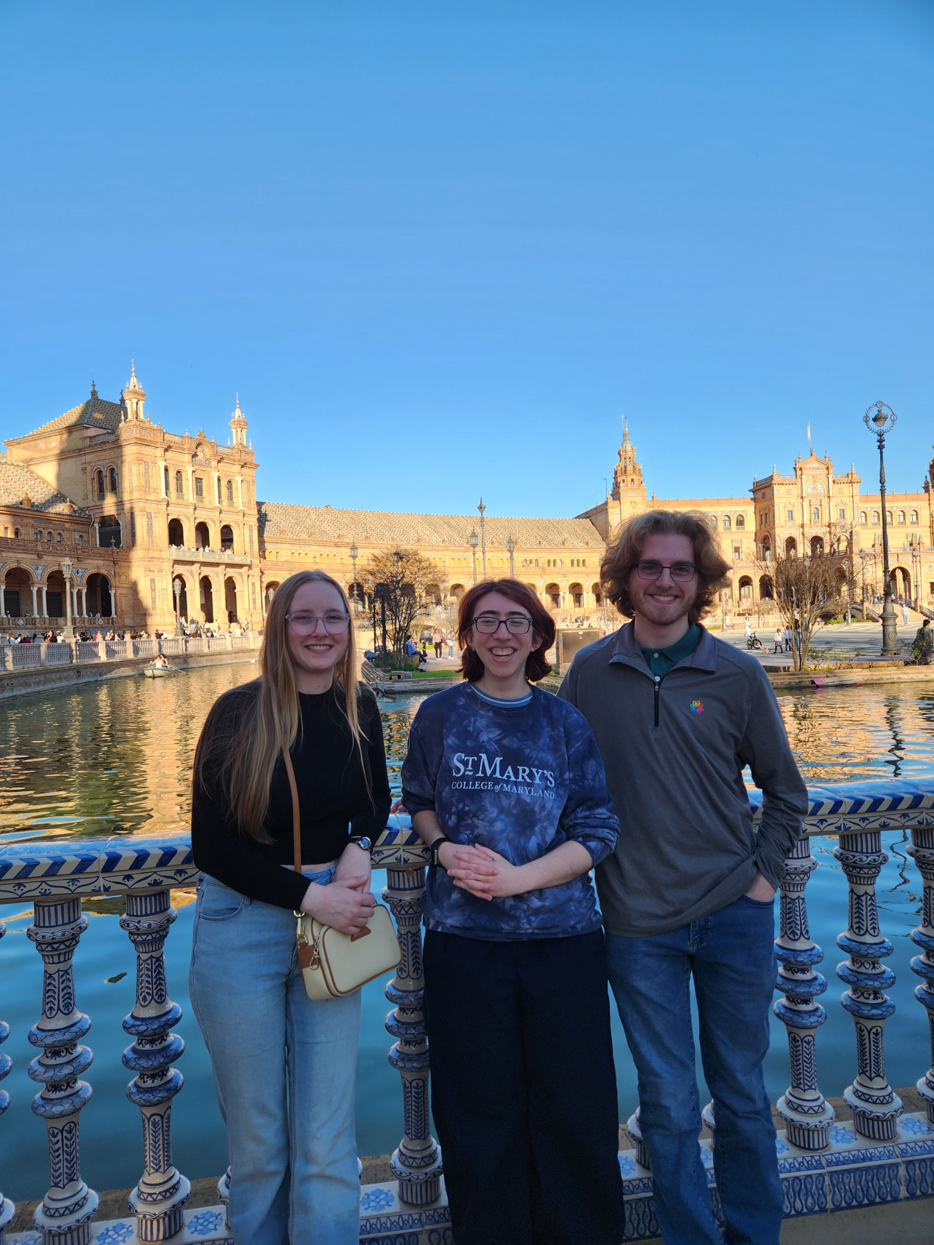 Three people stand smiling in front of a decorative railing at Plaza de España in Seville, Spain, with historic buildings and a canal in the background under a clear blue sky.