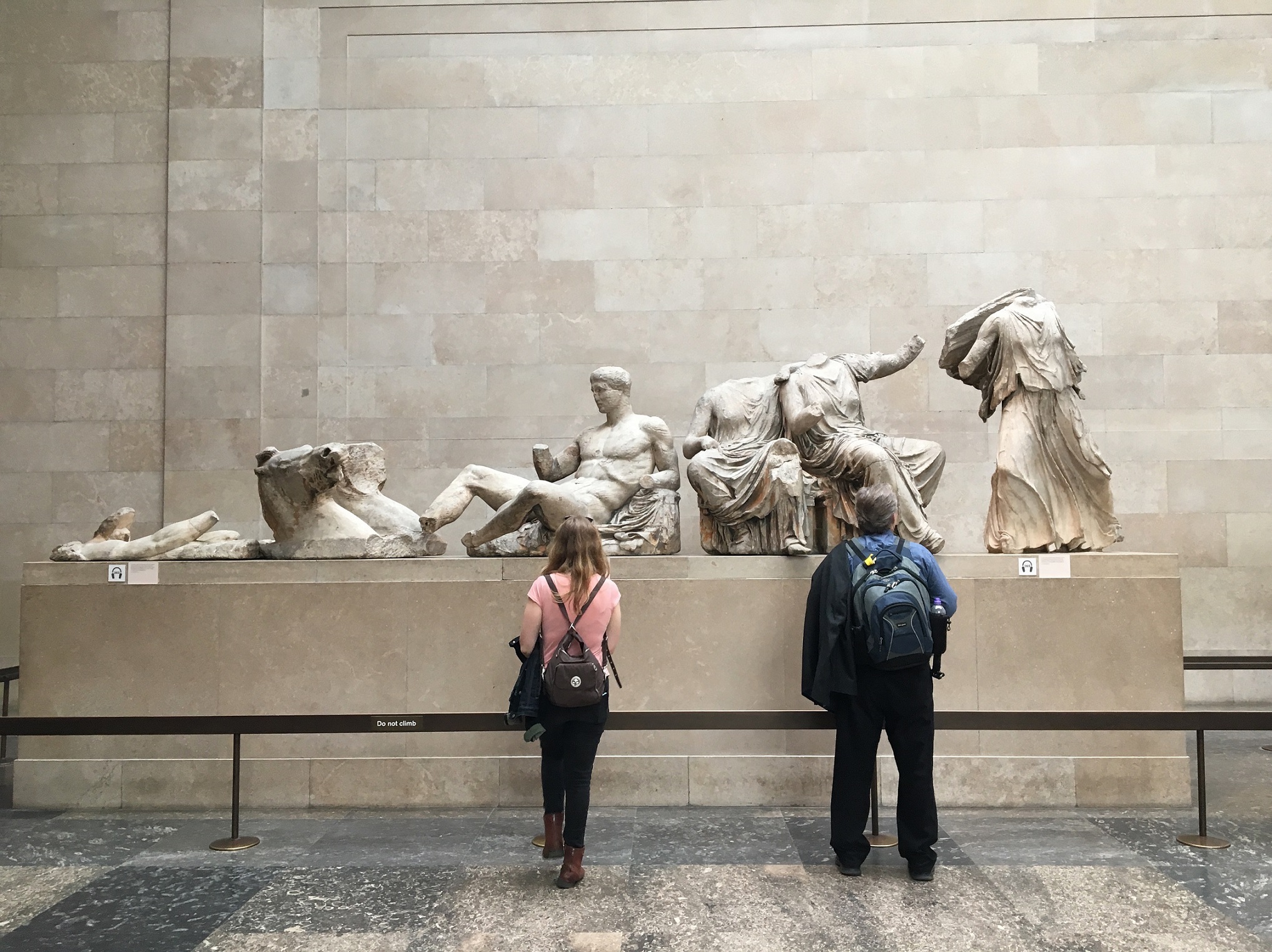 Two people stand in front of ancient marble Greek statues displayed on a plinth in a museum with a stone wall background.
