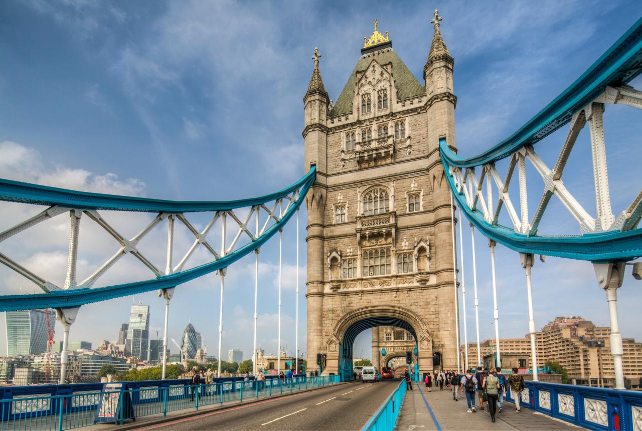 Tower Bridge in London, featuring its iconic towers and blue suspension elements, with pedestrians and vehicles crossing on a clear day.