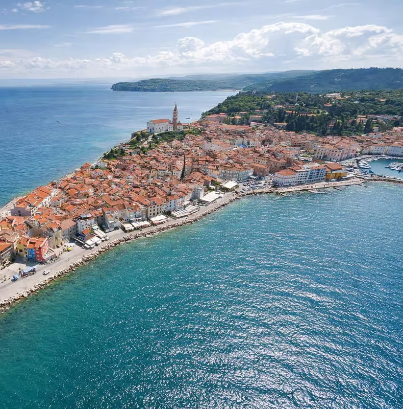 Aerial view of a coastal town with red-roofed buildings, a prominent church, and a curving shoreline bordered by blue sea under a partly cloudy sky.