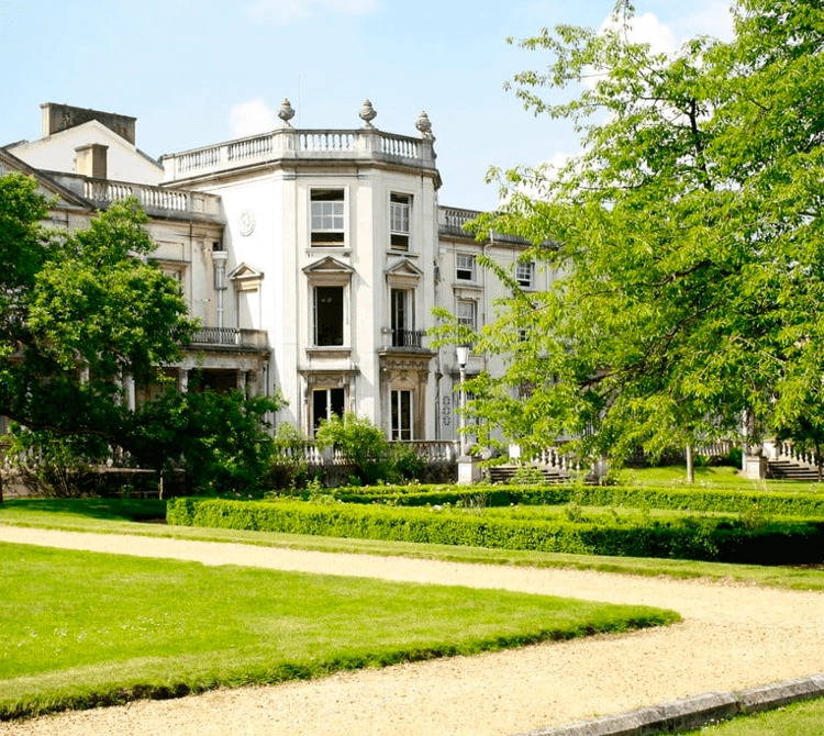 A large, white historic building with columns is surrounded by green lawns, trimmed hedges, and gravel pathways under a clear sky.