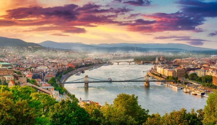A panoramic view of Budapest with the Danube River, historic buildings, bridges, and a colorful sunset sky.