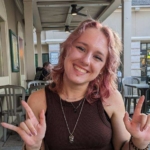 A person with shoulder-length, wavy, light pink hair smiles while sitting outdoors at a café, making hand signs with both hands.