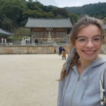 A woman in glasses and a gray hoodie smiles in front of a traditional Japanese temple with trees and hills in the background.