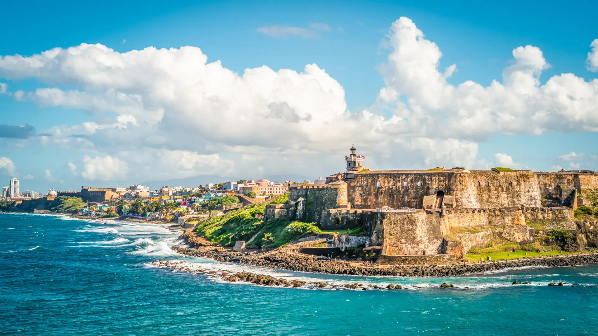 Historic stone fort overlooking the ocean with waves crashing against the rocky shore under a bright, partly cloudy sky.