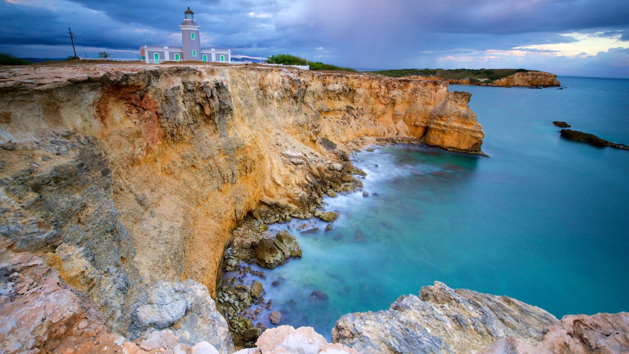A pastel green lighthouse sits atop rugged cliffs overlooking turquoise waters and rocky shores under a cloudy sky.