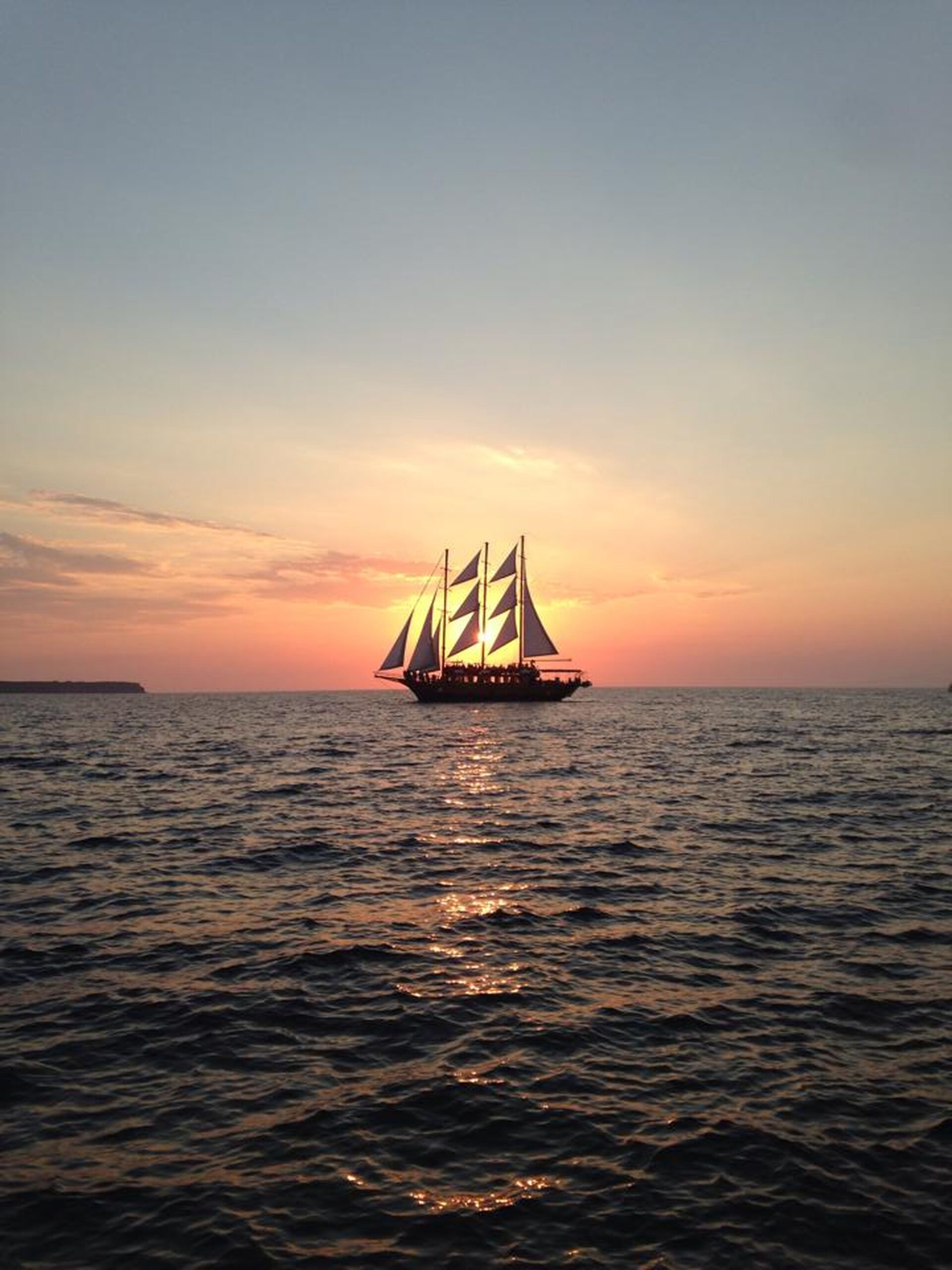 A sailing ship with raised sails moves across the sea at sunset, with the sky glowing orange and pink above the horizon.