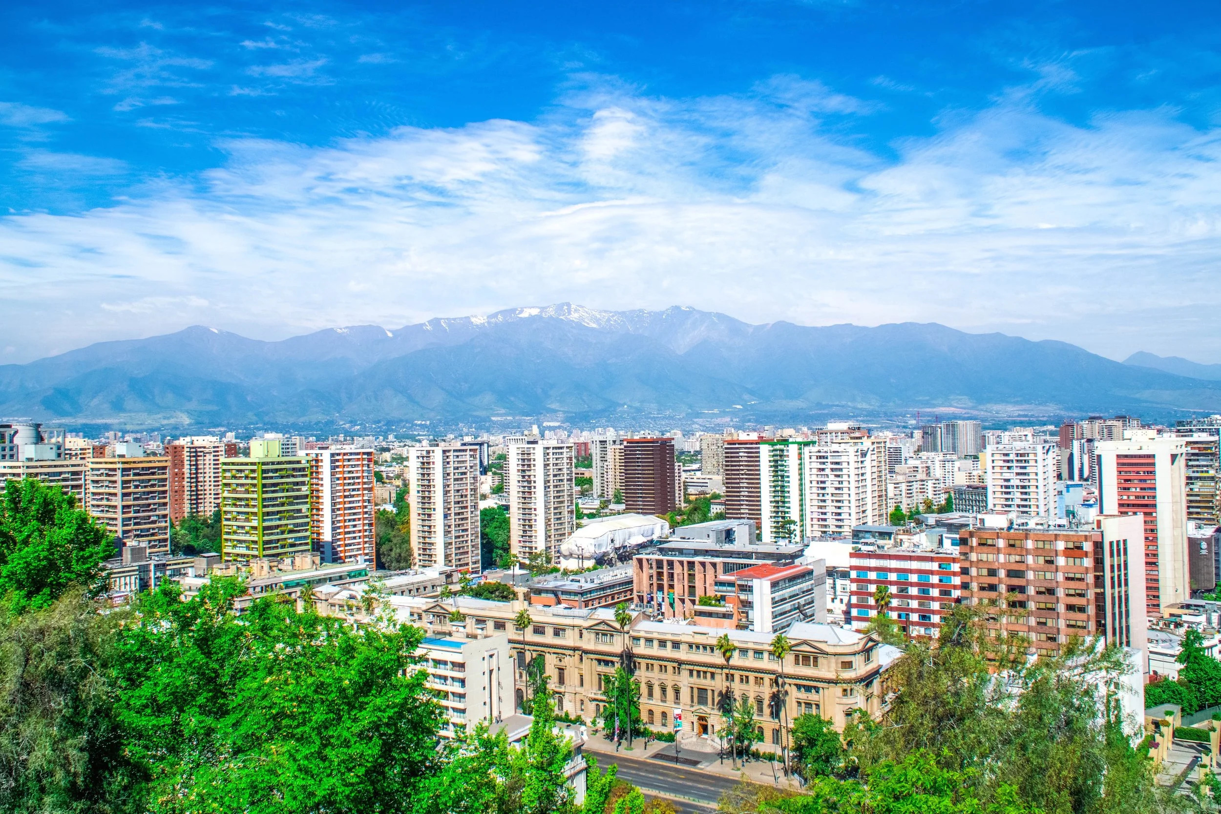 Cityscape with modern buildings, trees in the foreground, and mountains under a partly cloudy blue sky in the background.